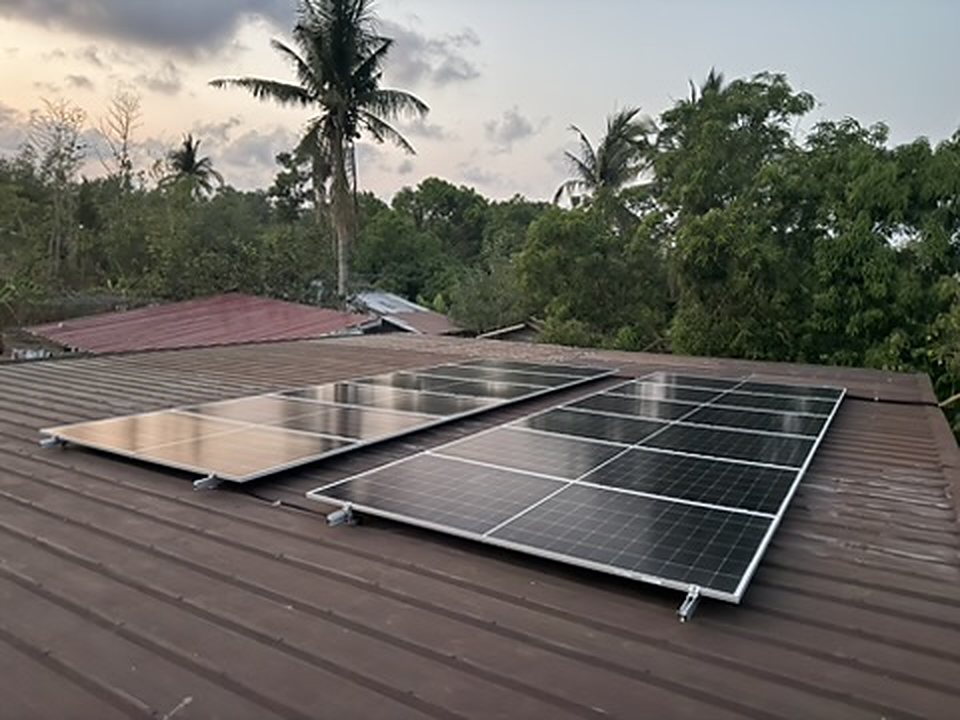 Two rows of solar panels on a provincial metal roof, with palm and mango trees behind